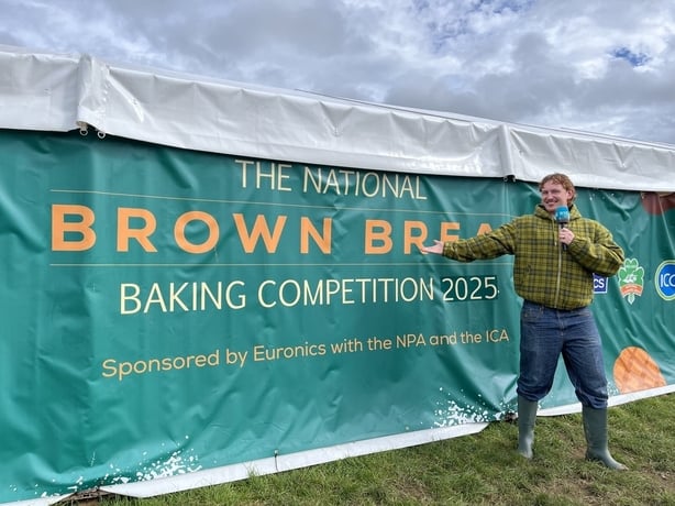 Pj Kirby standing in front of a green sign reading "The National Brown Bread Baking Competition"