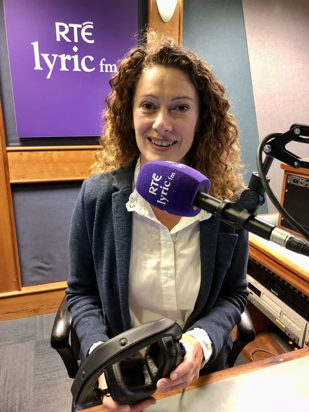 Anja Murray sits in a radio studio desk, holding headphones and smiling, with a microphone in front of her, with a purple mic shield saying RTE lyric fm in white letters. A big purple square is on the wall behind her with the same RTE lyric fm letters in white