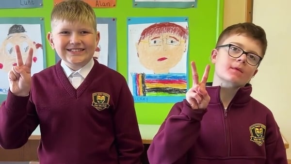 Two schoolkids in maroon uniforms in their classroom make the sign for the letter V in the Irish Sign Language alphabet.