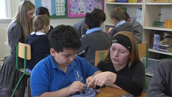 A young boy sitting at a table wearing a blue top is shown how to sew by a teacher in a classroom.