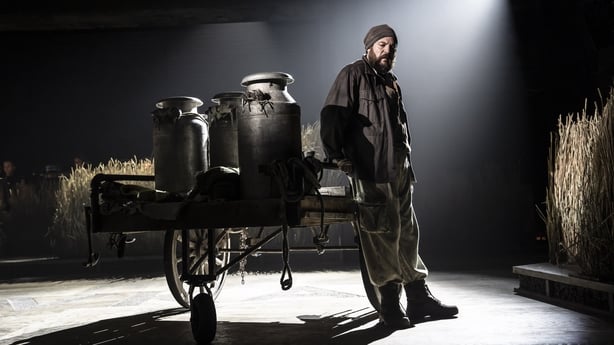 A photo on a stage of a man with a heavy beard leaning against an old milk cart with hay bales in the background