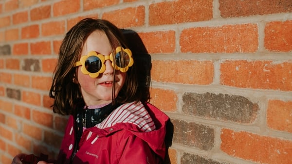 A child stands close to a red brick wall, casting a shadow. She wears novelty yellow flower shaped sunglasses. She looks off to the side. Conceptual with space for copy.