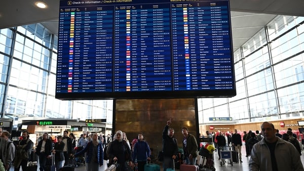 Image shows dozens of people looking at a large screen displaying flight departure and arrival times in an airport.