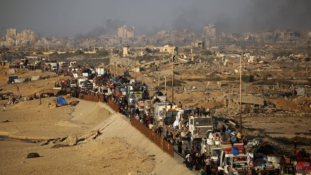 Image shows hundreds of people travelling by foot and car in a convoy along a dirt road with destroyed buildings in the background