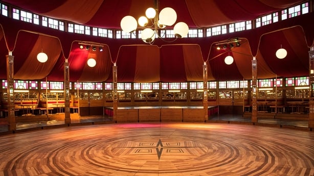 The inside of a large circular red tent with seating around the edges, looking on to a circular wooden floor and six bright circular lights hanging from the middle of the ceiling
