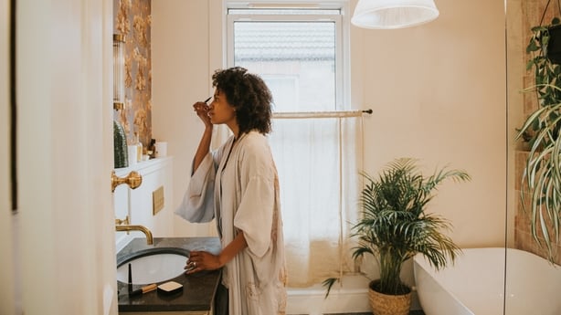 A beautiful black woman stands in a stylish bathroom in a long, silky robe and carefully applies her make-up.