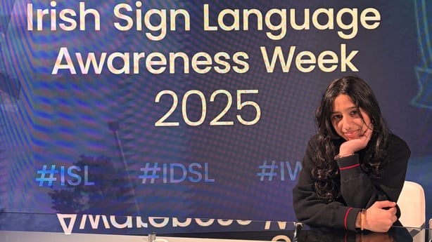 A young girl in a school uniform sits in front a screen with the words Irish Sign Language Awareness Week 2025