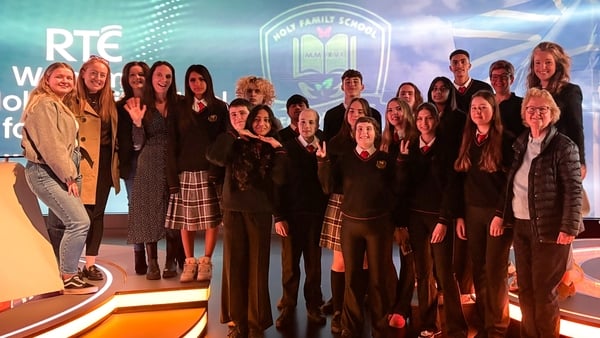 A group of TY students and adults pose in a TV studio with their school crest on a video wall in the background.