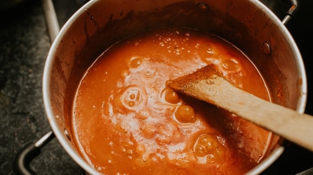 A bubbling pot of tomato sauce simmers on the stovetop, releasing savoury aromas throughout the kitchen. A wooden spoon stirs the sauce gently, highlighting the rich colour of the mixture.