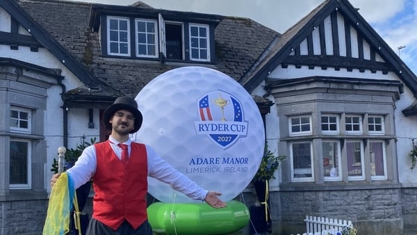 A man in a top hat and red vest is standing infront of a giant golf ball that is promoting the 2027 Ryder Cup that is being held in Adare Manor