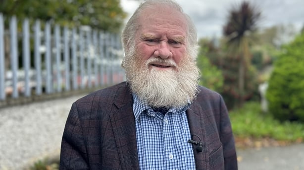 An elderly man with white hair and a white beard wears a blue shirt with a dark coloured suit jacket. He is standing in front of a white wall with grey railings on top. Trees and bushes are in the background.