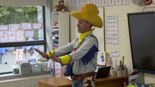 A man dressed as a cowboy wearing an oversized yellow hat gives a talk in a classroom.