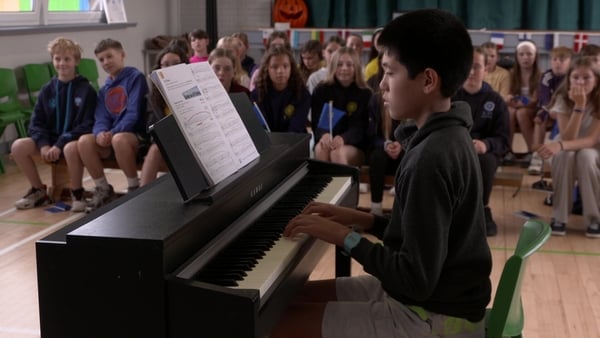 A yong boy in a black hoodie plays the piano in front of his class in a school hall.