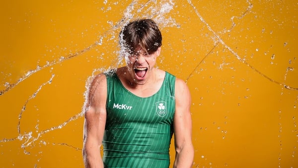 Fintan McCarthy in a green rowing suit pictured against an orange backdrop with water being thrown at him