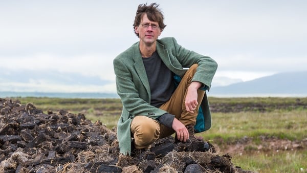 Manchán Magan crouches on top of a pile of turf outside in a field. He wears glasses, a long green coat and green jumper with beige trousers and his hair is tousled in the wind and he