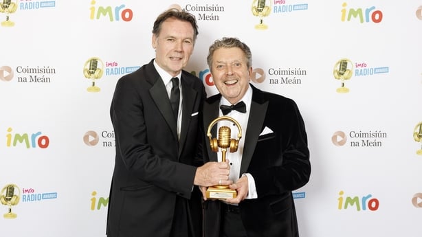 Michael O'Kane and George Hamilton dressed in suits and holding a gold trophy together