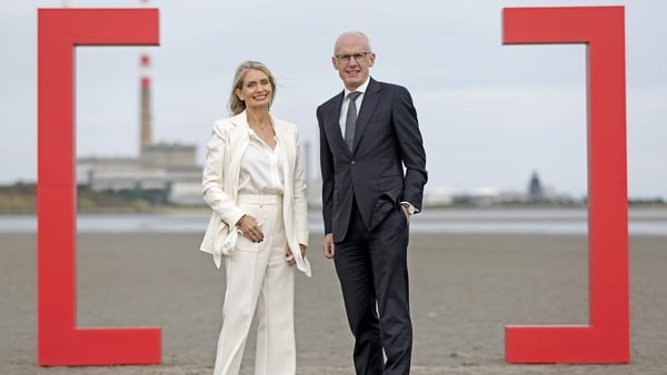 A businesswoman and a business man pose for photos on a beach with the Poolbeg chimneys behind them