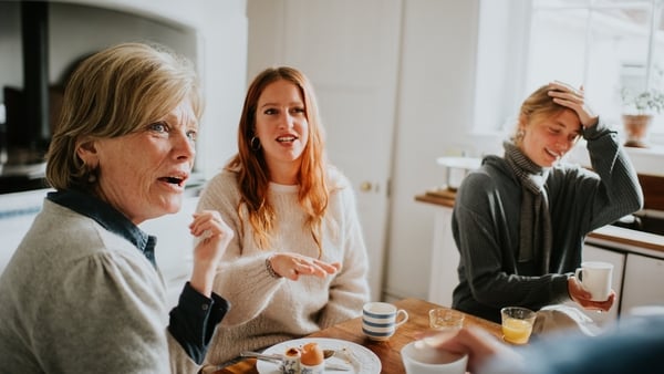 A family has an animated discussion in a kitchen. They are passionate about their opinions, talking over one another. One of the family members in the background looks frustrated, refusing to engage.