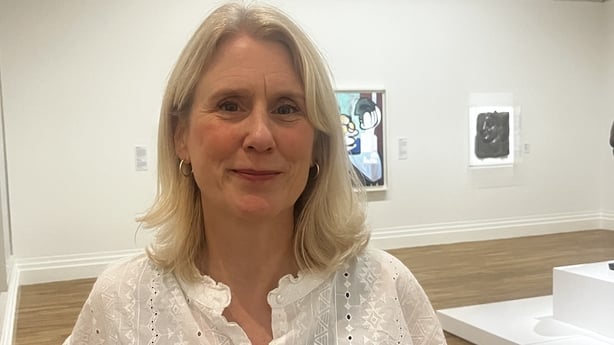 A woman with shoulder length blonde hair is standing in front of a white wall with a colourful painting on it. She is wearing a white lacey blouse.