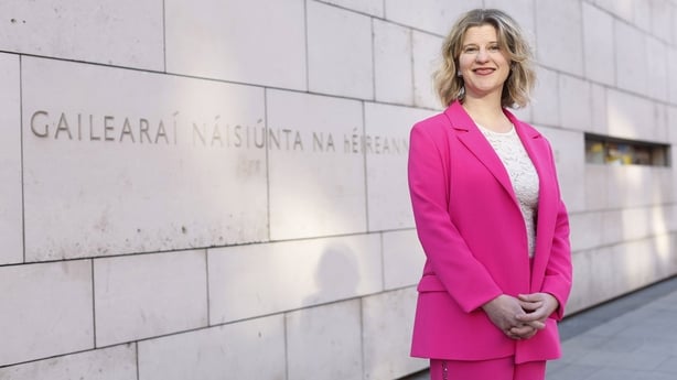 A woman with shoulder length blonde hair is wearing a bright pink suit with a white blouse. She is standing with her hands clasped in front of a pale grey stone building.