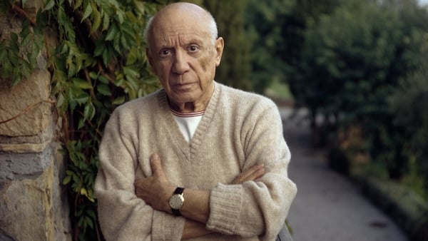 An older man white hair is wearing a beige jumper. His arms are folded. He is standing on a path with trees and hedges behind him.