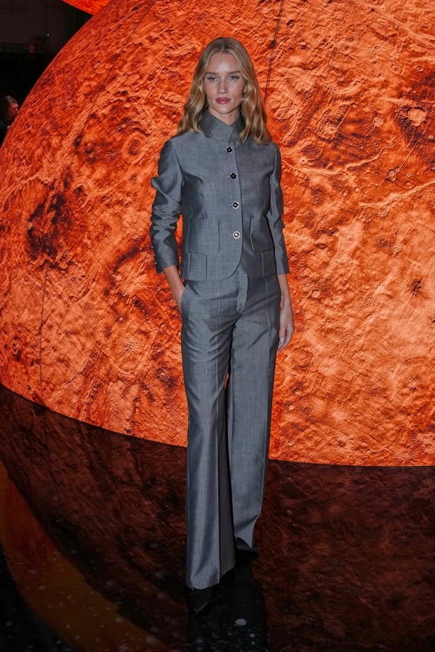 Rosie Huntington-Whiteley poses in a grey suit in front of a large globe shaped like the sun