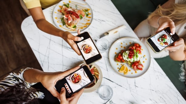 From a top-down perspective, the hands of three women can be seen using smartphones to photograph colourful dishes at a cafe