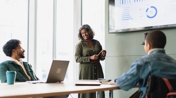 Worker stands at front of meeting room presenting to colleagues, holding pencil to point at different parts of presentation and smiling at male colleague