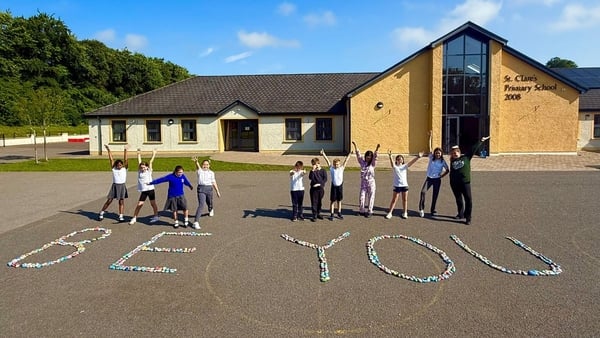 A group of children stand behind a sign made out of pebbles which reads 'be you'