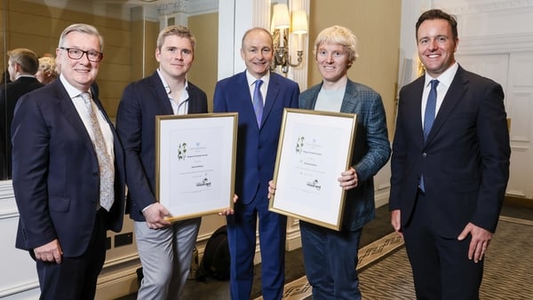 A group of five men in suits with two of them holding awards