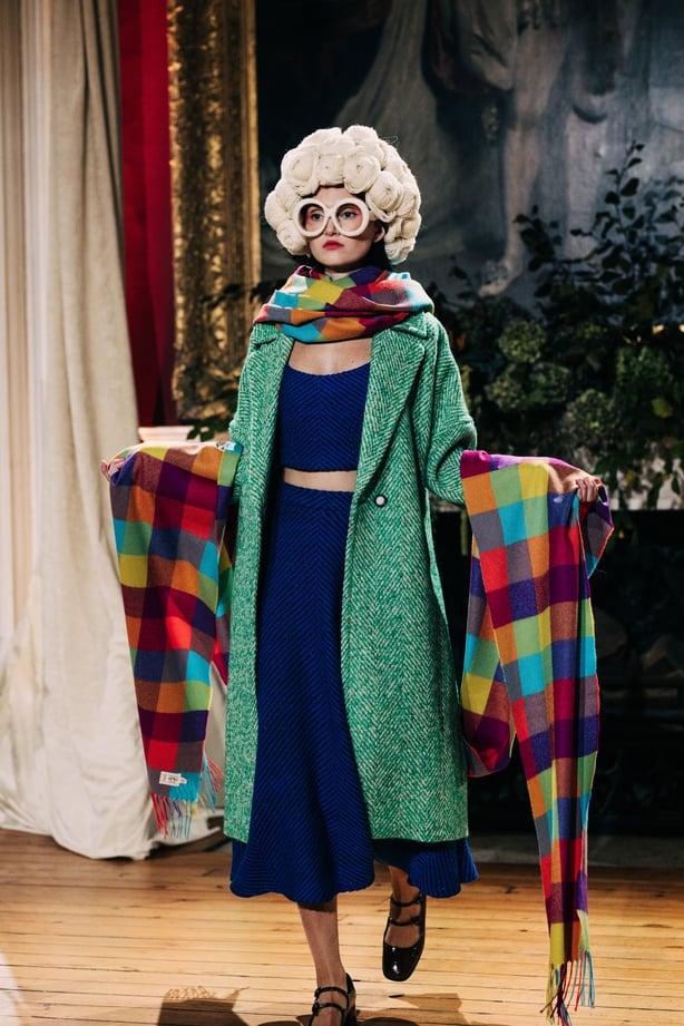 A model wering a green coat, blue top and skirt and a multicoloured scarf