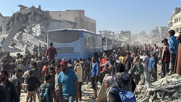 Dozens of men and boys are following a convoy of buses along a road. Rubble and damaged buildings can be seen in the background.