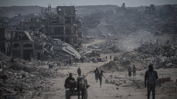 People move along dusty roads with destroyed buildings in the background