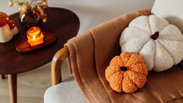 Home interior with cozy burning candle, dry flower decor on table and soft pumpkin-shaped pillows and warm orange plaid on chair in the living room. Home comfort, coziness, aromatherapy.