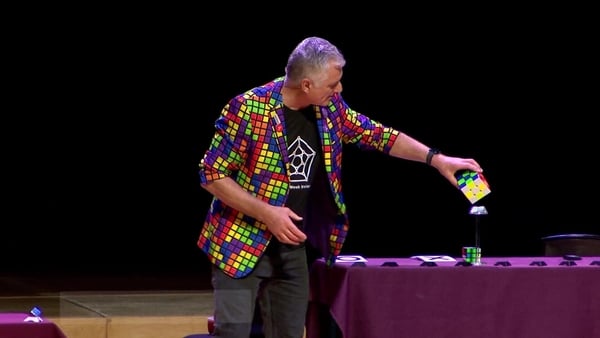 A man in a multi coloured jacket on stage shows off a rubiks cube trick.