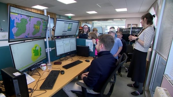 Meteorologists at Met Éireann show how they can forecast weather. One man is sitting in front of 4 screens while a woman explains to the children standing in the background.