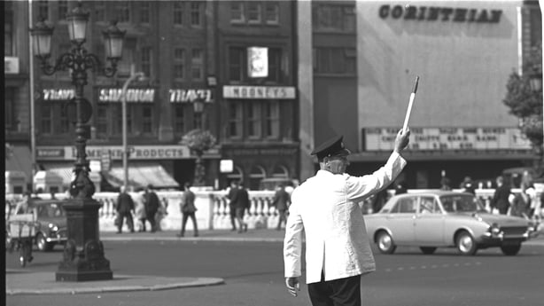 A traffic control garda on duty at O'Connell Bridge, Dublin on 30 July 1969. He is standing in right profile with his baton raised. He is wearing a hat and gloves. There are two cars on the bridge; pedestrians can be seen walking on the footpath beside it. Some of the buildings along Eden Quay can b