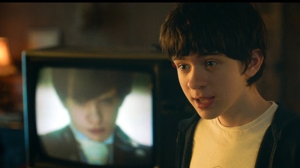 A close-up of a young boy with an identical version of him on a TV screen in the background.