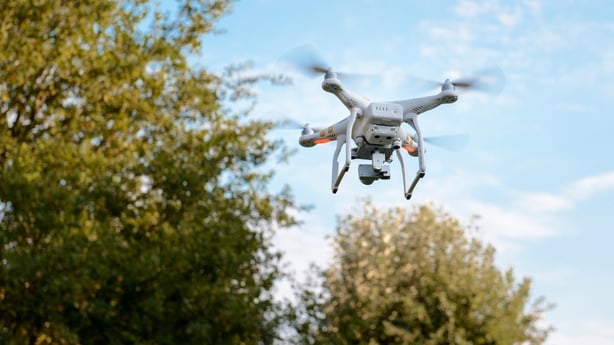 A grey coloured drone flies over green tries with a blue sky backdrop.