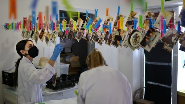 Dozens of colourful pegs are pinning damaged photographs to a line of string as a dark haired woman in a white lab coat looks at them.