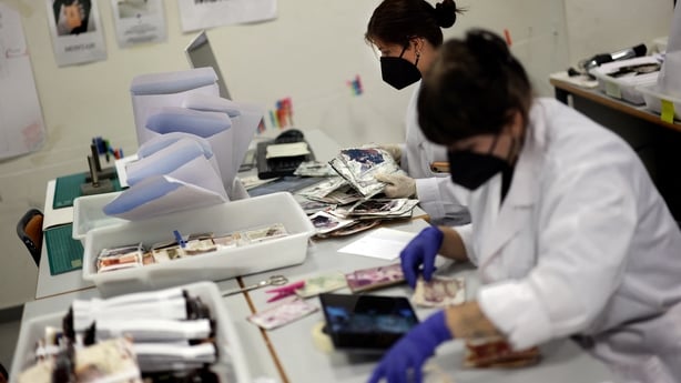Two dark haired women wearing black face masks and white lab coats sit at a desk sorting through damaged photographs.