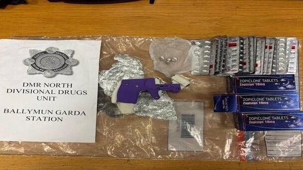Image shows a small purple handgun lying on a brown table beside a clear plastic packet with an assortment of tablets of various sizes and colours.