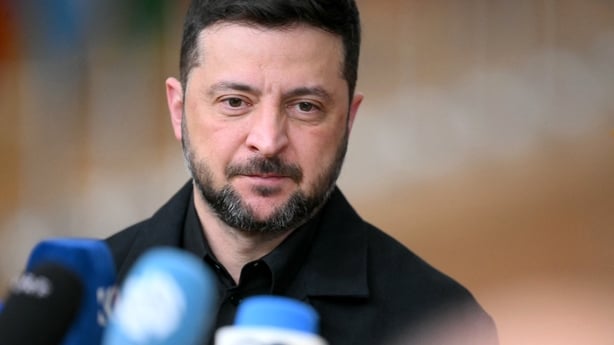 A close up head and shoulders image of Ukrainian President Volodymyr Zelensky. He has short, dark hair and a trimmed beard. He is wearing a dark coloured shirt and is standing in front of three blue microphones.