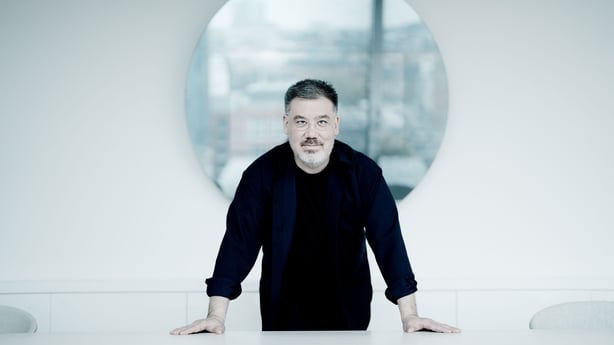 Alan Gilbert is standing wearing all black and leaning slightly forward on a white surface. The background is white with a circular shaped image behind him. It's slightly blurred, it could be a landscape or even a window looking out onto a landscape