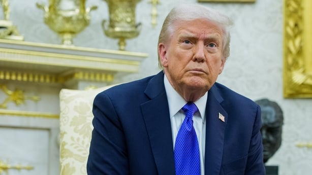 A close up head and shoulders image of US President Donald Trump. He is looking above the camera lense. He is wearing a white shirt, bright blue tie and navy jacket. A white and gold fireplace can be seen in the background.