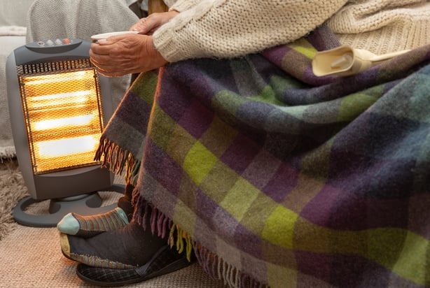 Elderly pensioner trying to keep warm near an electric heater with blanket and a warm drink.
