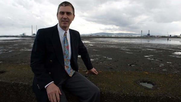 A business man in a suit sitting at a waterfront with industrial chimneys in the background