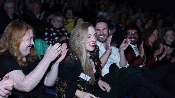 writer Lynda McCarthy rises from the audience to take 1st prize at the 2025 RTÉ Short Story Competition, surrounded by clapping onlookers at awards night in Pavilion Theatre Dun Laoghaire