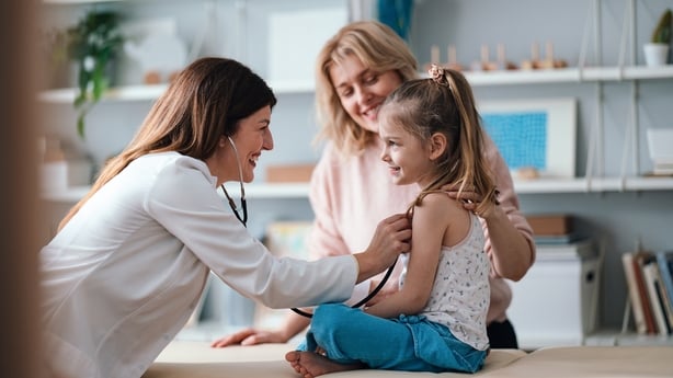 A friendly pediatrician performs a health examination for a happy young girl, observed by her caring mother. The image conveys family care, child health, and professional medical service.