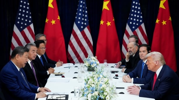 US President Donald Trump, wearing a white shirt, red tie and dark suit, sits at a table with several other men, all wearing white shirts, dark suits and red or blue ties. There are white and blue flowers on the table. There are red and yellow Chinese flags, and blue, white and red US flags in the b